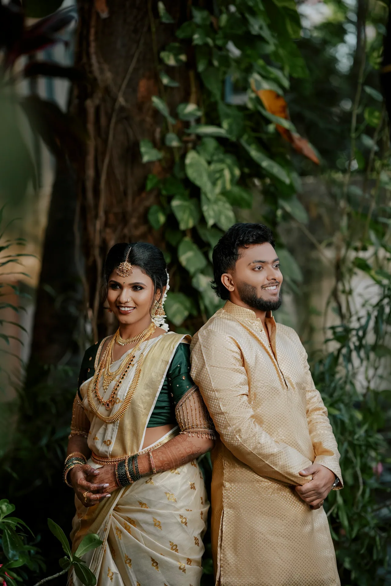 Wedding couple back to back by a tree, the bride in a cream-and-gold kasavu saree and the groom in a gold kurta, Chennai — LNC Photography.